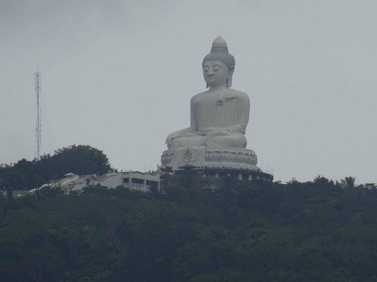 The white Big Buddha statue on the forested ridge of Nakkerd Hill, Phuket, seen from Wat Chalong under a grey sky