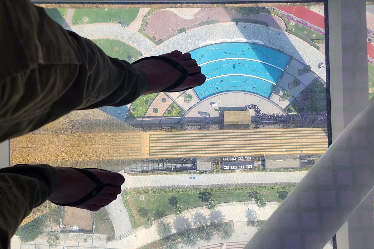 Visitor's feet on the glass floor panel of the Dubai Frame bridge, looking straight down at Zabeel Park and the fountain far below