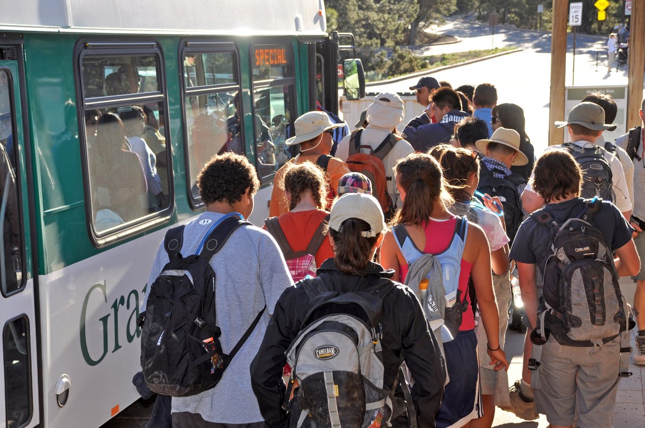 Visitors with backpacks lining up to board a white and green Grand Canyon shuttle bus on the South Rim