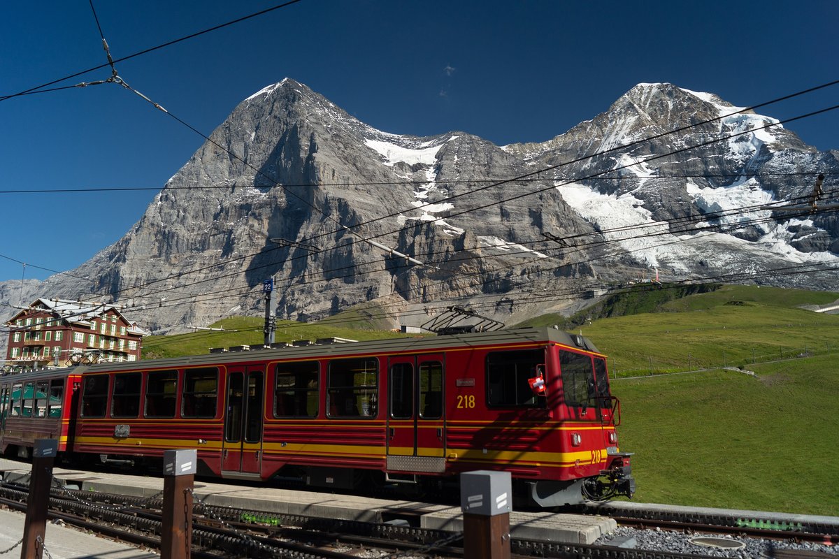 A red and yellow Jungfrau Railway train at Kleine Scheidegg station, with the Eiger north face and the Mönch behind it under a clear blue sky