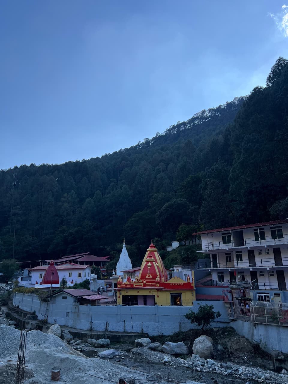 Neem Karoli Baba Ashrams in India: Kainchi, Vrindavan, Lucknow and More Kainchi Dham ashram with red-and-yellow and white temple spires and ashram buildings below a forested hill, with the rocky riverbed in front