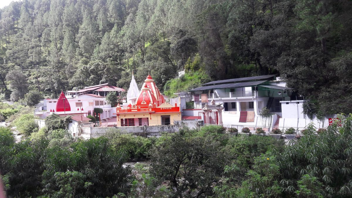Jim Corbett to Kainchi Dham Distance: Route via Nainital, Taxi Fare and Bus Options Red and white temple spires and white ashram buildings below a pine-forested hillside at Kainchi Dham, Nainital district