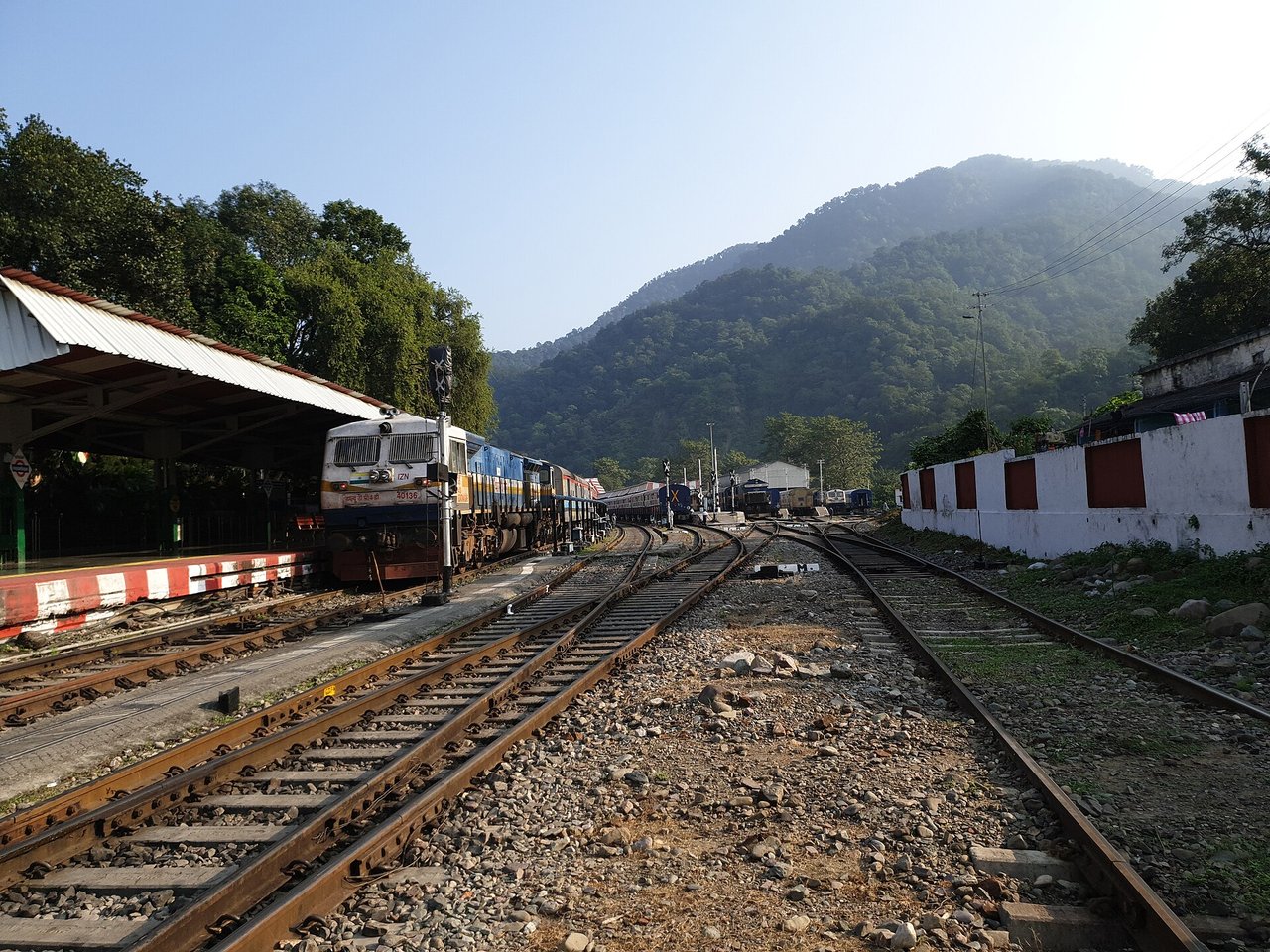 Haridwar to Kainchi Dham Distance: Train + Taxi Route and Travel Time Railway tracks and a diesel locomotive at Kathgodam railway station with forested hills behind under a clear sky
