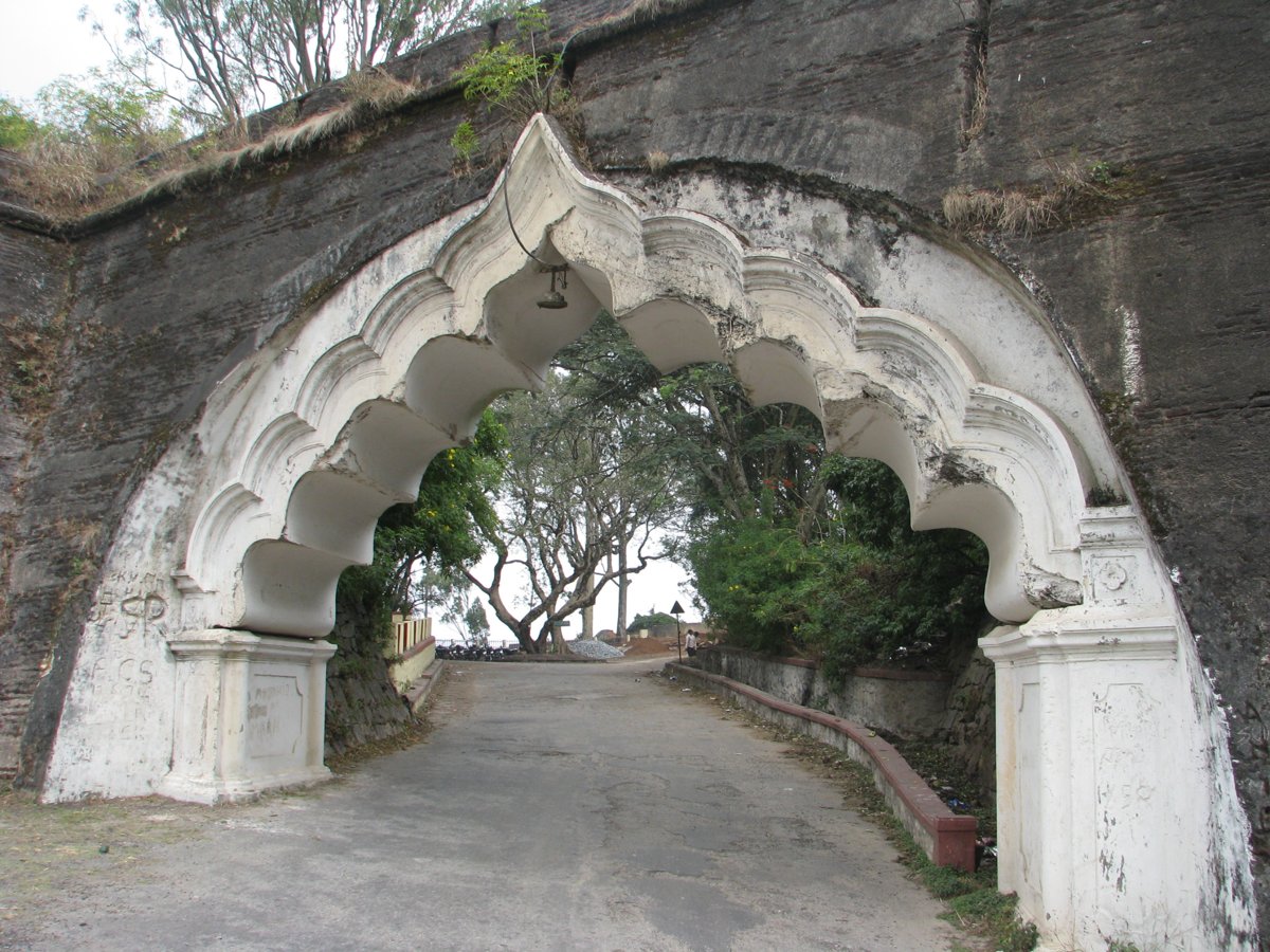 Nandi Hills Entry Fee, Timings, Parking Charges and Sunrise Tips White arched gateway set in the old dark stone fort wall on Nandi Hills, with a paved road passing through under trees