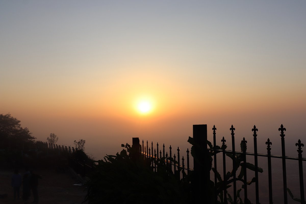 Nandi Hills Entry Fee, Timings, Parking Charges and Sunrise Tips Orange sun rising in a hazy sky above the plains, seen past a dark iron railing and plants at a Nandi Hills viewpoint