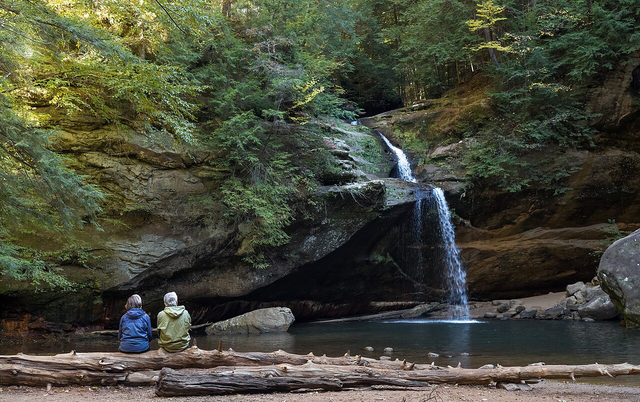 Two visitors sitting on a log beside a waterfall and pool in the Old Man's Cave area of Hocking Hills State Park in autumn