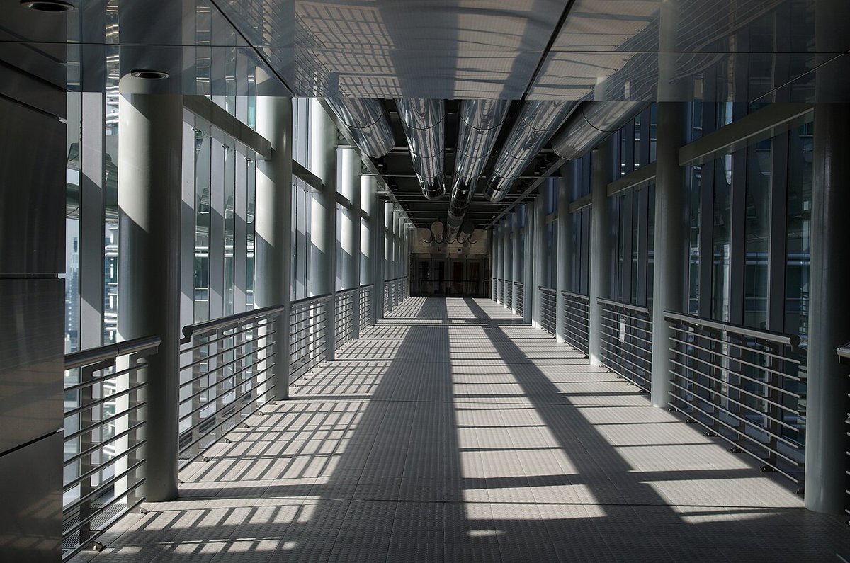 Inside the Petronas Twin Towers Skybridge: a long glass-walled walkway with steel railings and sunlight casting stripes on the floor