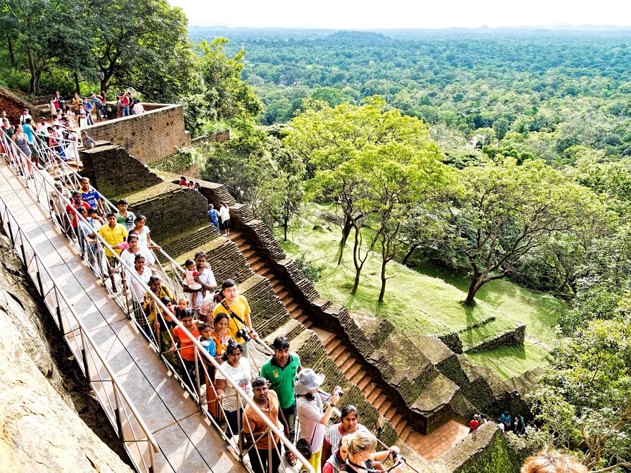 Sigiriya Entrance Fee for Foreigners, SAARC (India) Visitors and Children Visitors queuing on a railed stone walkway and stairway on the side of Sigiriya rock, with forest and gardens far below