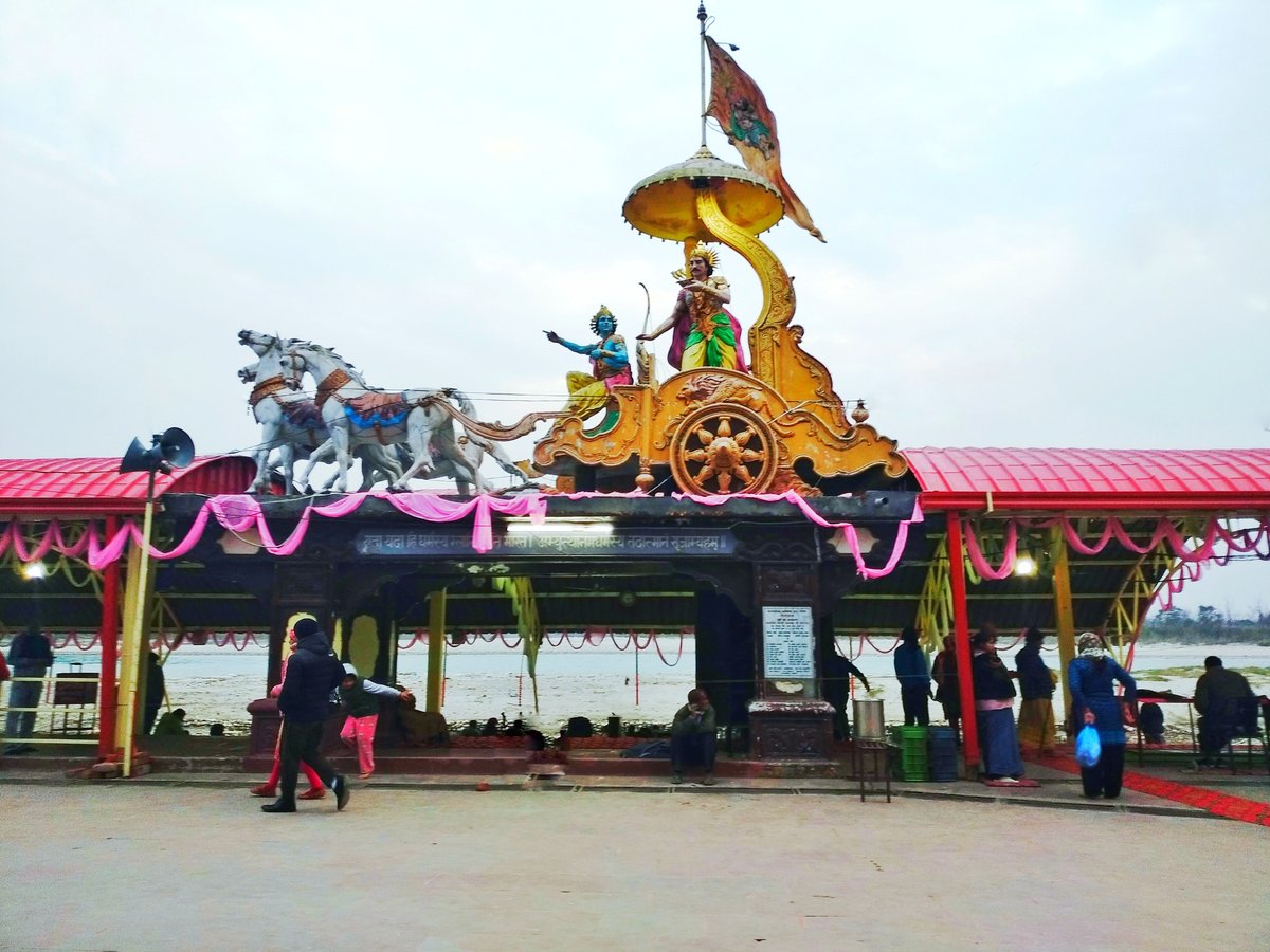 Painted chariot statue of Krishna and Arjuna with white horses above a pavilion at Triveni Ghat, Rishikesh, with the Ganga behind