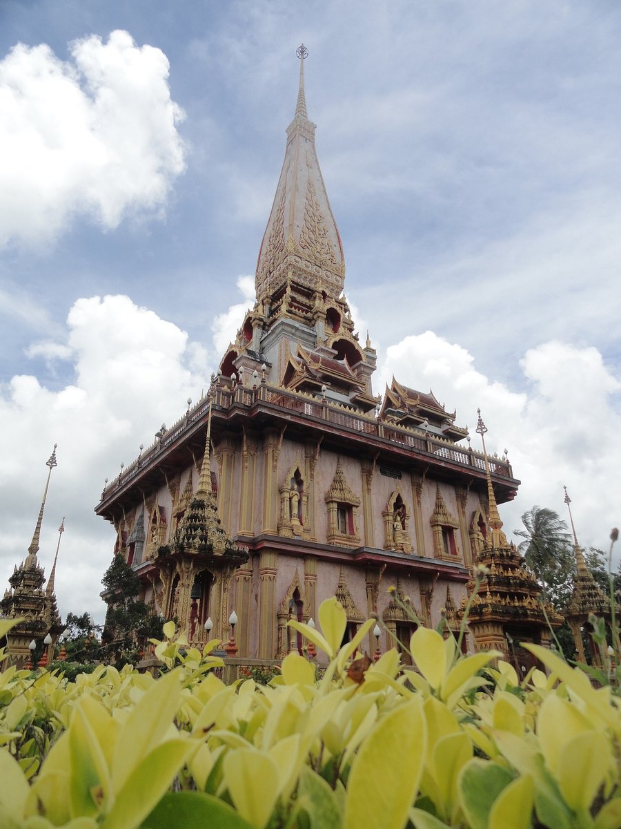 Wat Chalong Phuket: Opening Hours, Dress Code and What to See The tall grand chedi of Wat Chalong in Phuket, with a pointed spire, a terrace balcony and gold decoration, seen behind a hedge under a cloudy sky
