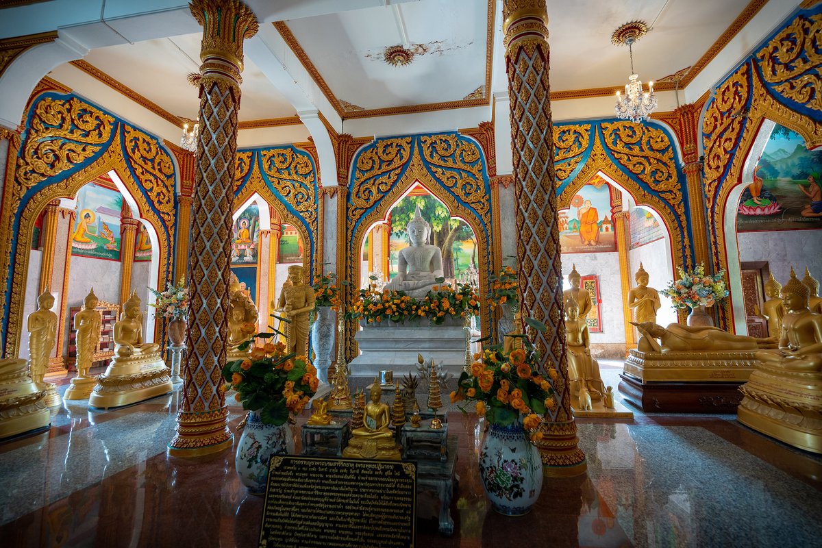 Wat Chalong Phuket: Opening Hours, Dress Code and What to See Gilded Buddha images, carved pillars and colourful murals inside a hall at Wat Chalong, Phuket, with a white seated Buddha at the centre
