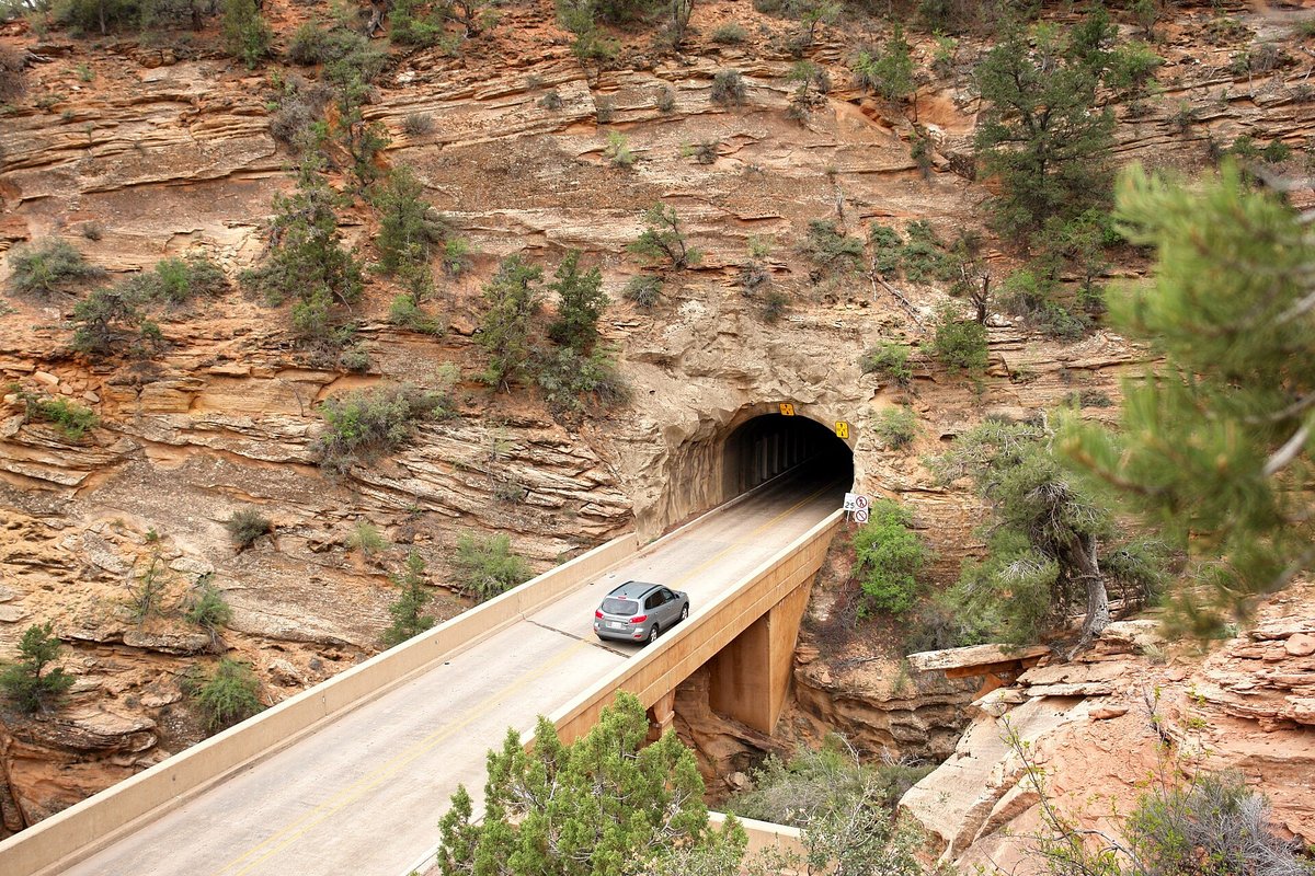 A silver car on a concrete bridge heading out of a tunnel portal cut into layered sandstone on the Zion-Mount Carmel Highway